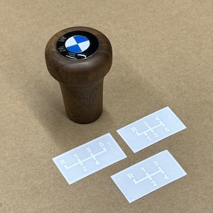 Puede incluir: Una palanca de cambios de madera con un logotipo BMW azul y blanco en la parte superior. Se muestran tres pegatinas blancas con patrones de cambio de marchas sobre una superficie marr&oacute;n.