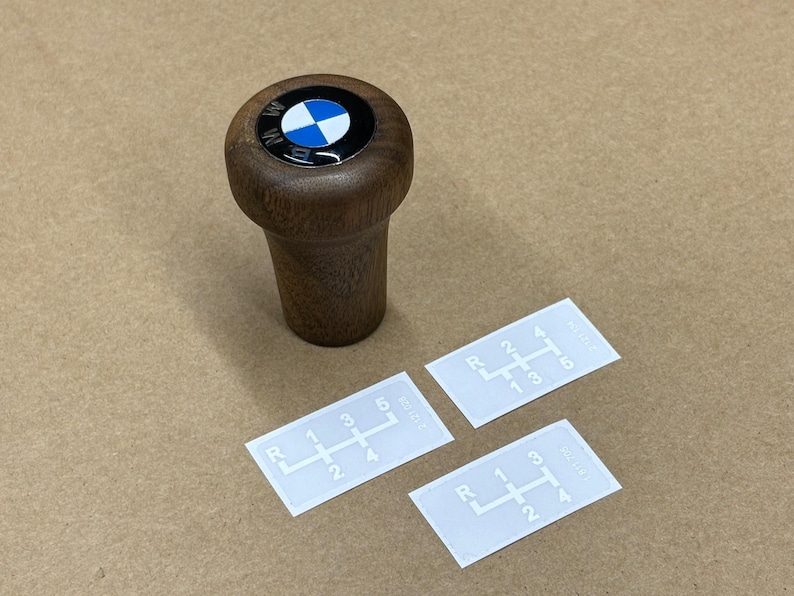 Puede incluir: Una palanca de cambios de madera con un logotipo BMW azul y blanco en la parte superior. Se muestran tres pegatinas blancas con patrones de cambio de marchas sobre una superficie marr&oacute;n.