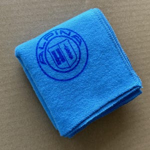 May include: A folded, square, bright blue microfiber cloth. A circular logo in blue is printed on the cloth. The logo has the word "ALPINA" in a circle around a shield design.