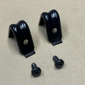 May include: Two black metal brackets with a curved top and a mounting hole, plus two black screws. The brackets have a right-angle design and a small rectangular base.