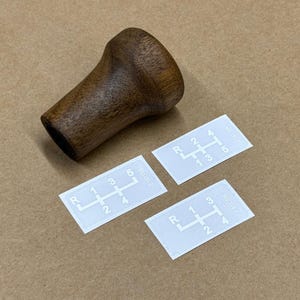 Puede incluir: Tres pegatinas blancas con n&uacute;meros y letras negras que muestran diferentes patrones de cambio de marchas. Las pegatinas est&aacute;n sobre una superficie marr&oacute;n junto a una palanca de cambios de madera marr&oacute;n.