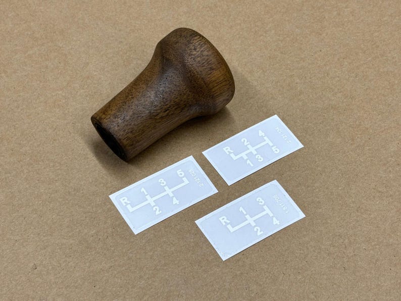 Puede incluir: Tres pegatinas blancas con n&uacute;meros y letras negras que muestran diferentes patrones de cambio de marchas. Las pegatinas est&aacute;n sobre una superficie marr&oacute;n junto a una palanca de cambios de madera marr&oacute;n.