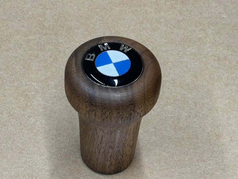 Puede incluir: Una palanca de cambios de madera con un logotipo BMW negro y azul en la parte superior.