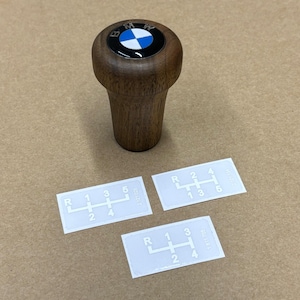 Puede incluir: Una palanca de cambios de madera con un logotipo BMW azul y blanco en la parte superior. Tres pegatinas blancas con patrones de cambio de marchas se muestran sobre una superficie marr&oacute;n.