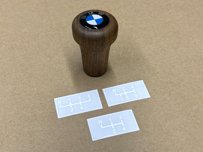 Puede incluir: Una palanca de cambios de madera con un logotipo BMW azul y blanco en la parte superior. Tres pegatinas blancas con patrones de cambio de marchas se muestran sobre una superficie marr&oacute;n.