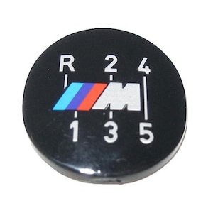 May include: A black gear shift knob with white numbers and letters indicating the gear positions: R, 1, 2, 3, 4, and 5. The BMW logo is in the center, featuring blue, red, and light blue stripes. The knob is round.