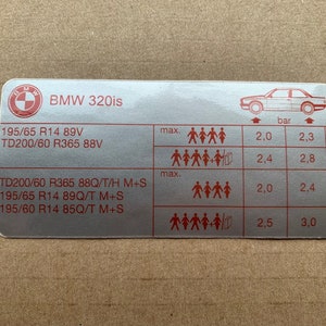 Può includere: Un adesivo argentato con il testo "BMW 320is" e un diagramma che mostra le dimensioni degli pneumatici e il carico massimo e la pressione corrispondenti in bar. Le dimensioni degli pneumatici indicate sono 195/65 R14 89V, TD200/60 R365 88V, TD200/60 R365 88Q/T/H M+S, 195/65 R14 89Q/T M+S e 195/60 R14 85Q/T M+S.