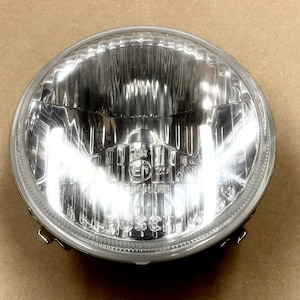 May include: A round, clear glass headlight with a chrome rim. The headlight has a ridged lens and is marked with the brand name "BOSCH" and other markings. The headlight is designed for automotive use.