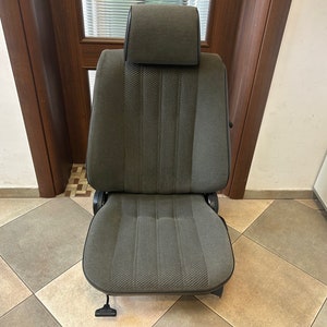 May include: A gray car seat with a headrest. The seat has a textured fabric with vertical stripes and a dark gray trim. The seat is designed for a vehicle and has a lever on the bottom.