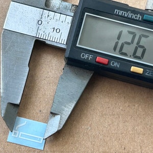 May include: A silver caliper measuring a small, light blue rectangle with a white outline of a cigarette. The caliper reads 12.7 mm.