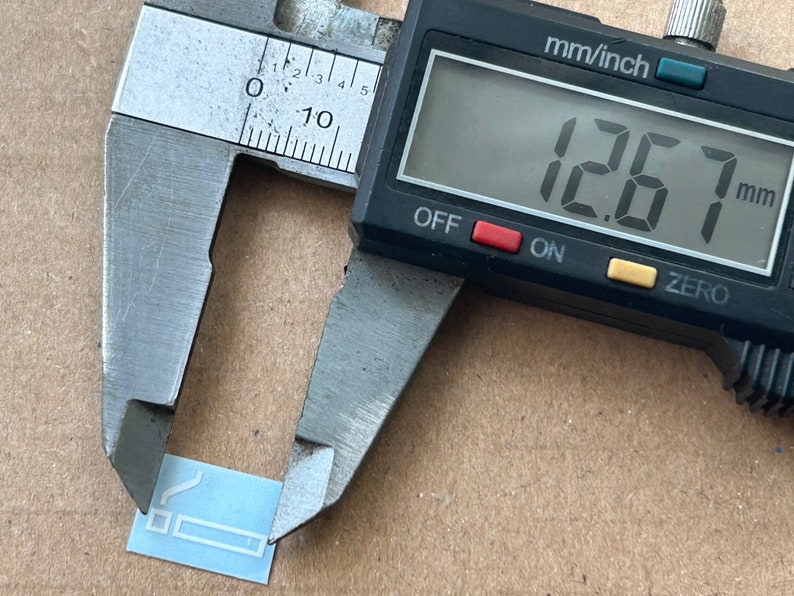 May include: A silver caliper measuring a small, light blue rectangle with a white outline of a cigarette. The caliper reads 12.7 mm.