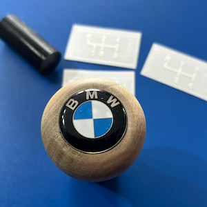 Puede incluir: Una palanca de cambios de madera con un logotipo BMW negro y azul. El logotipo tiene las letras "BMW" en letras plateadas alrededor del diseño de hélice azul y blanco.