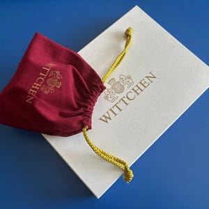 May include: A white box with the text "WITTCHEN" in gold lettering. The box is partially covered by a red velvet drawstring bag with the text "WITTCHEN" in gold lettering.