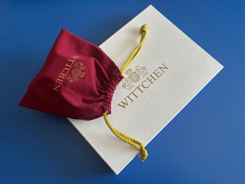 May include: A white box with the text "WITTCHEN" in gold lettering. The box is partially covered by a red velvet drawstring bag with the text "WITTCHEN" in gold lettering.