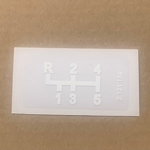 Könnte beinhalten: Ein weißer Aufkleber mit einem Schaltmuster, der die Gänge R, 1, 2, 3, 4 und 5 anzeigt. Auf dem Aufkleber ist auch die Nummer 2 121 134 aufgedruckt. Der Aufkleber ist rechteckig.