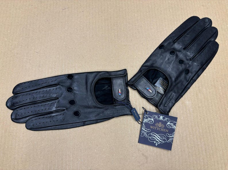 May include: A pair of black leather driving gloves with a perforated design and a small logo tag that reads "WITTCHEN".