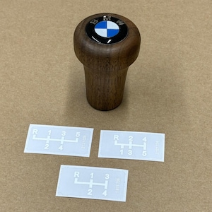 Puede incluir: Tres pegatinas blancas con n&uacute;meros y letras impresos en negro que muestran un patr&oacute;n de cambio de marchas para un autom&oacute;vil. Las pegatinas est&aacute;n sobre una superficie marr&oacute;n. Tambi&eacute;n hay un pomo de cambio de marchas de madera con un logotipo BMW negro y azul sobre la superficie.