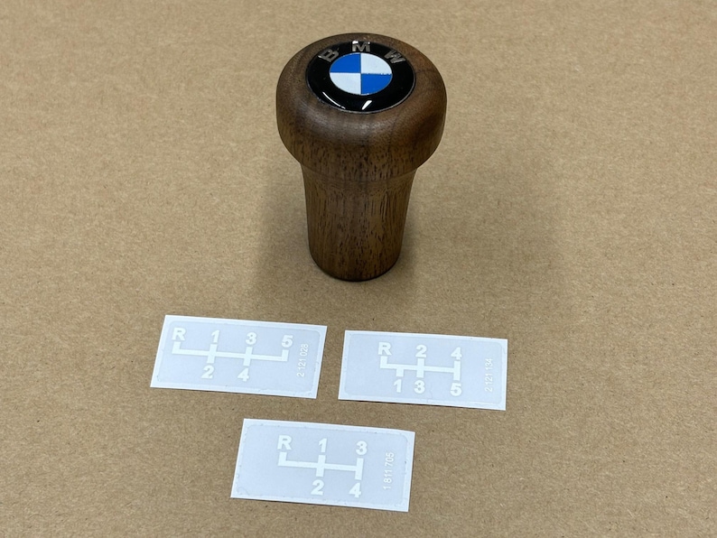 Puede incluir: Tres pegatinas blancas con n&uacute;meros y letras impresos en negro que muestran un patr&oacute;n de cambio de marchas para un autom&oacute;vil. Las pegatinas est&aacute;n sobre una superficie marr&oacute;n. Tambi&eacute;n hay un pomo de cambio de marchas de madera con un logotipo BMW negro y azul sobre la superficie.