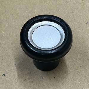 Könnte beinhalten: Ein schwarzer und silberner Knopf mit einer runden, flachen Oberseite und einem zylindrischen Sockel.