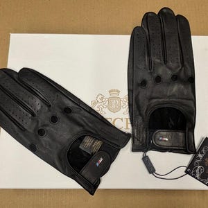 May include: Two pairs of black leather driving gloves with a perforated design. The gloves have a wrist strap and a small logo on the strap.