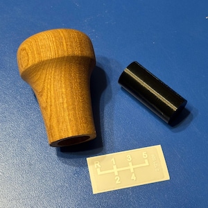 May include: A white ceramic mug with a black handle and the text "But First, Coffee" in a bold, black font. The mug is set against a blurred background, suggesting a cozy setting.