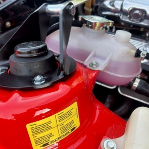 Puede incluir: Primer plano del compartimento del motor de un coche rojo con una pieza de metal negra y plateada, un depósito de refrigerante de plástico transparente con líquido rosa y una etiqueta de advertencia amarilla que dice "Hidráulica: Al utilizar un elevador hidráulico, asegúrese de que el vehículo esté correctamente asegurado antes de elevarlo o bajarlo. No exceda la capacidad de elevación máxima del elevador. Asegúrese de que el elevador esté correctamente mantenido e inspeccionado regularmente.  Automático: En caso de fallo hidráulico, el vehículo se bajará automáticamente al suelo.  El sistema hidráulico está diseñado para proporcionar una experiencia de elevación segura y fiable.  Alpine"