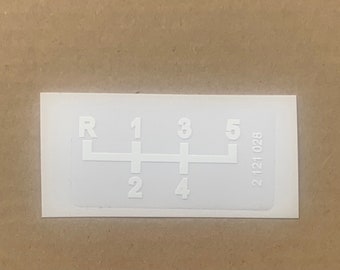 May include: A white sticker featuring a gear shift pattern with the letters R, and numbers 1, 2, 3, 4, and 5. The sticker also has the number 2 121 028 printed on the right side.