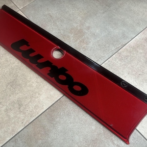 Puede incluir: Panel rojo con superficie texturizada y la palabra "turbo" en letras negras. El panel tiene un borde negro y un agujero circular. Pieza de automóvil.