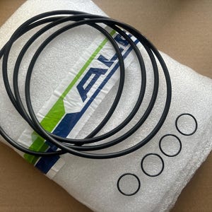 May include: A collection of black rubber O-rings of varying sizes. Several larger rings are looped together, while smaller individual rings are arranged below. The items rest on a white foam surface with a green and blue striped label.