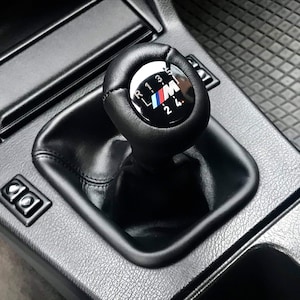 Peut inclure: Pommeau de levier de vitesses en cuir noir avec une garniture argentée et un logo BMW noir et bleu avec les chiffres 1, 2, 3, 4, R et M.