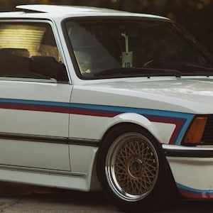 Könnte beinhalten: Ein weißer BMW 3er mit blauen, roten und weißen Rennstreifen. Das Auto hat ein Schiebedach und goldene Felgen.