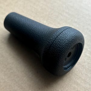 Puede incluir: Un pomo negro texturizado con un mango cilíndrico y una base redondeada y ensanchada. La superficie tiene una textura granulada. El pomo está diseñado para una aplicación mecánica o automotriz.