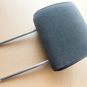 May include: A gray car headrest with a rectangular cushion and two chrome support rods. The headrest is made of a textured fabric and has a black leather trim around the edges. The rods are straight and have rounded tips.