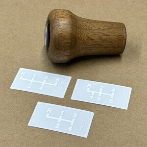 Puede incluir: Tres pegatinas blancas con n&uacute;meros y letras negras que muestran un patr&oacute;n de cambio de marchas de 5 velocidades. Las pegatinas est&aacute;n sobre una superficie marr&oacute;n junto a una perilla de cambio de marchas de madera.