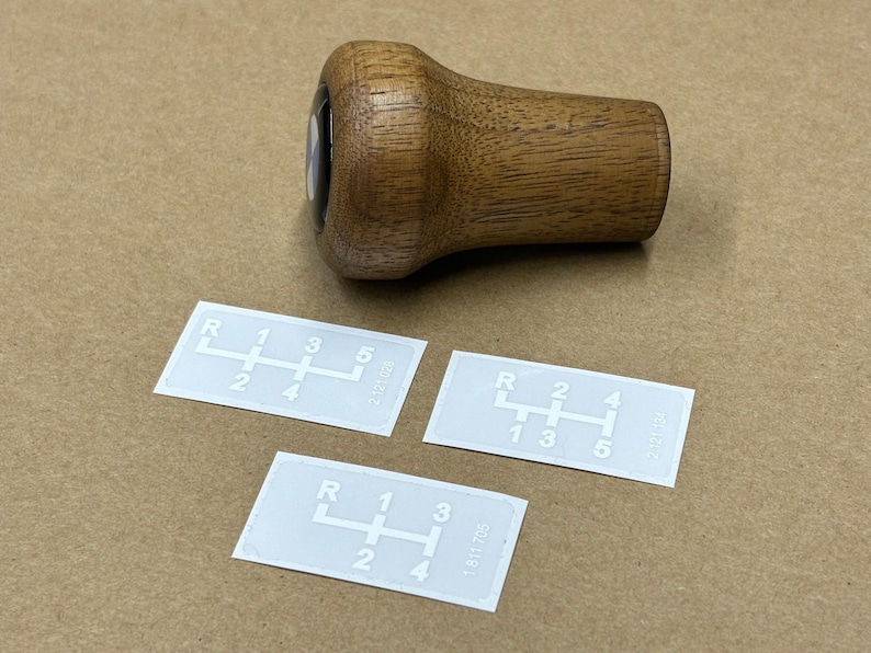 Puede incluir: Tres pegatinas blancas con n&uacute;meros y letras negras que muestran un patr&oacute;n de cambio de marchas de 5 velocidades. Las pegatinas est&aacute;n sobre una superficie marr&oacute;n junto a una perilla de cambio de marchas de madera.