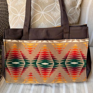 May include: A brown tote bag with a geometric patterned front panel. The pattern features red, orange, yellow, and green diamond shapes on a tan background. The bag has brown handles and a brown top.