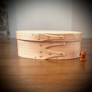 May include: A wooden oval box with a hinged lid and copper accents. The box is made of light wood and has a smooth finish.