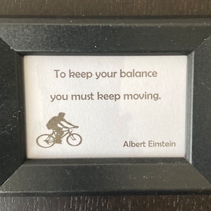 May include: A framed print with a black frame. The print features the quote "To keep your balance you must keep moving" by Albert Einstein, along with a silhouette of a person riding a bicycle.
