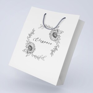 May include: White paper shopping bag with a black ink illustration of a floral wreath featuring sunflowers and the word "Elegance" in cursive.