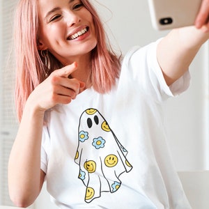 May include: White t-shirt featuring a ghost design with yellow smiley faces and blue flowers. The ghost has black eyes and a smiling mouth. The shirt is being held up by a person with pink hair.