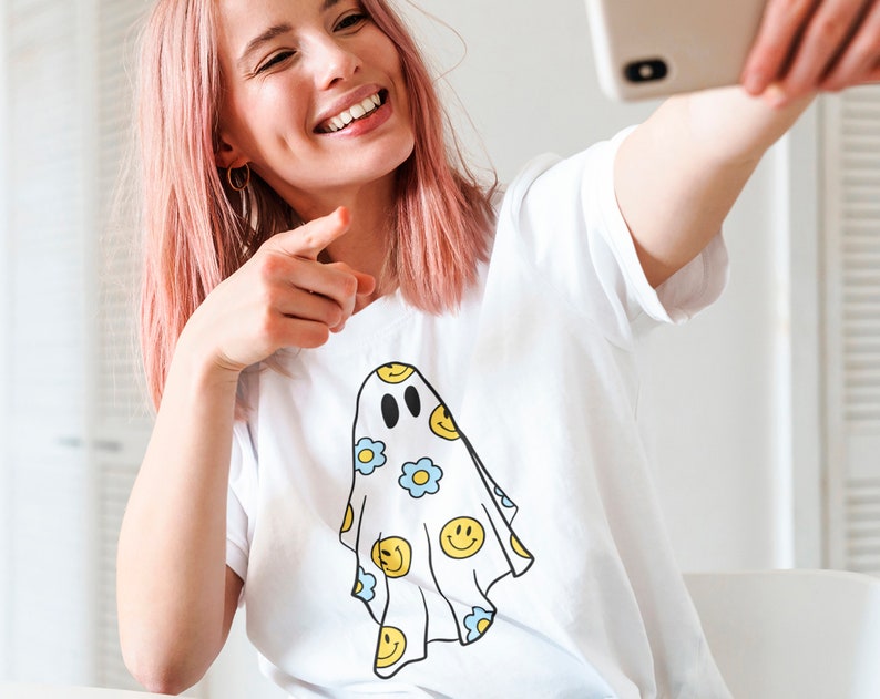 May include: White t-shirt featuring a ghost design with yellow smiley faces and blue flowers. The ghost has black eyes and a smiling mouth. The shirt is being held up by a person with pink hair.