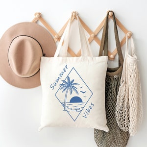 Pu&ograve; includere: Una borsa tote bianca con una grafica blu di una palma, il sole e le onde dell'oceano. Il testo "Summer Vibes" &egrave; scritto in un diamante blu attorno alla grafica.