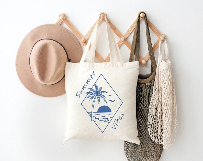 Pu&ograve; includere: Una borsa tote bianca con una grafica blu di una palma, il sole e le onde dell'oceano. Il testo "Summer Vibes" &egrave; scritto in un diamante blu attorno alla grafica.