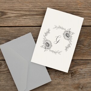May include: A white card with a black and white floral wreath design. The wreath is made of sunflowers and leaves. The letter "G" is in the centre of the wreath.