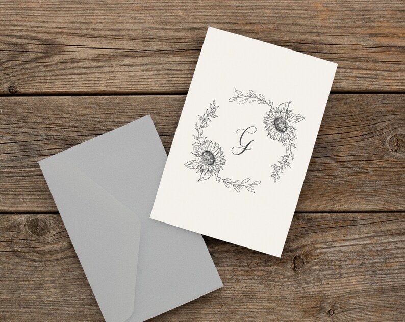 May include: A white card with a black and white floral wreath design. The wreath is made of sunflowers and leaves. The letter "G" is in the centre of the wreath.