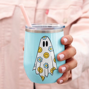 May include: A light blue insulated tumbler with a silver rim and lid. The tumbler features a white ghost design adorned with yellow smiley faces and blue flowers. A pink straw is inserted into the lid.