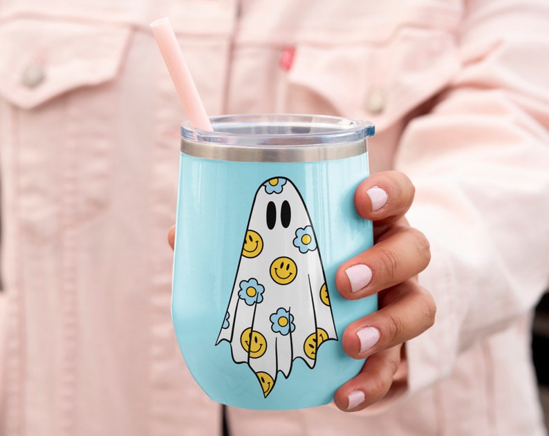 May include: A light blue insulated tumbler with a silver rim and lid. The tumbler features a white ghost design adorned with yellow smiley faces and blue flowers. A pink straw is inserted into the lid.