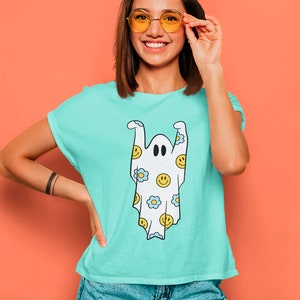 May include: A light blue t-shirt featuring a cartoon ghost design. The ghost is white with black eyes and is decorated with yellow smiley faces and blue flowers. The shirt has short sleeves and a crew neck.