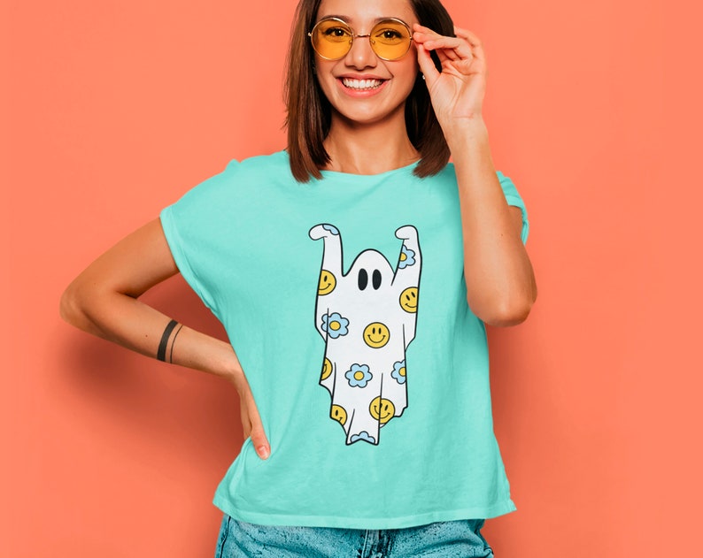 May include: A light blue t-shirt featuring a cartoon ghost design. The ghost is white with black eyes and is decorated with yellow smiley faces and blue flowers. The shirt has short sleeves and a crew neck.