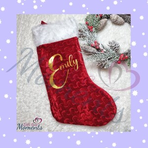 Può includere: Una calza di Natale rossa con un bordo in finta pelliccia bianca e il nome "Emily" in oro. La calza ha un motivo strutturato ed è decorata con una piccola ghirlanda con bacche rosse e pigne.